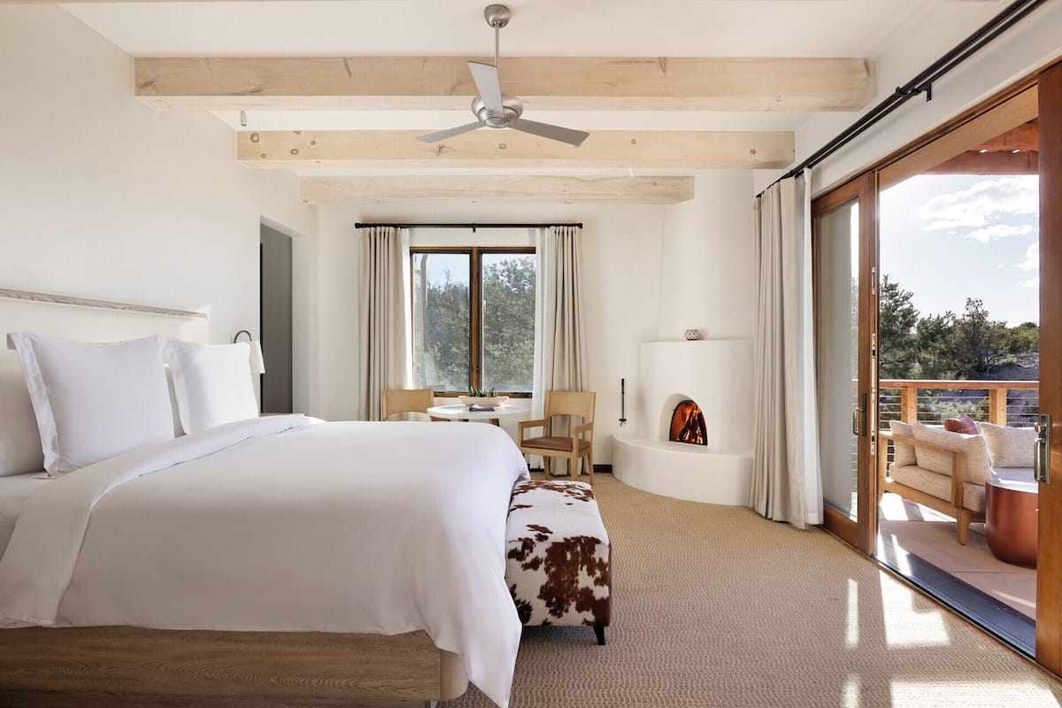 A bedroom with a large bed, a desk, a chair, a fireplace, and a balcony with outdoor furniture.