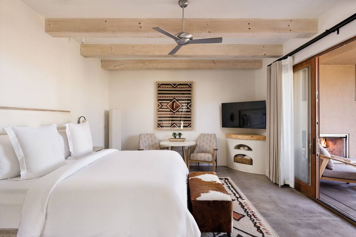 A bedroom with a bed, a ceiling fan, a TV, a small table with a plant, and a sliding glass door to a balcony.
