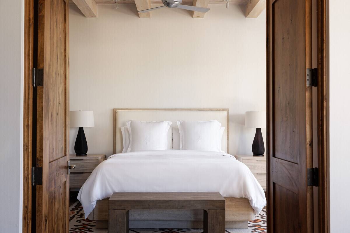 A bedroom with a wooden headboard, white bedding, two bedside tables with lamps, and a bench at the foot of the bed.