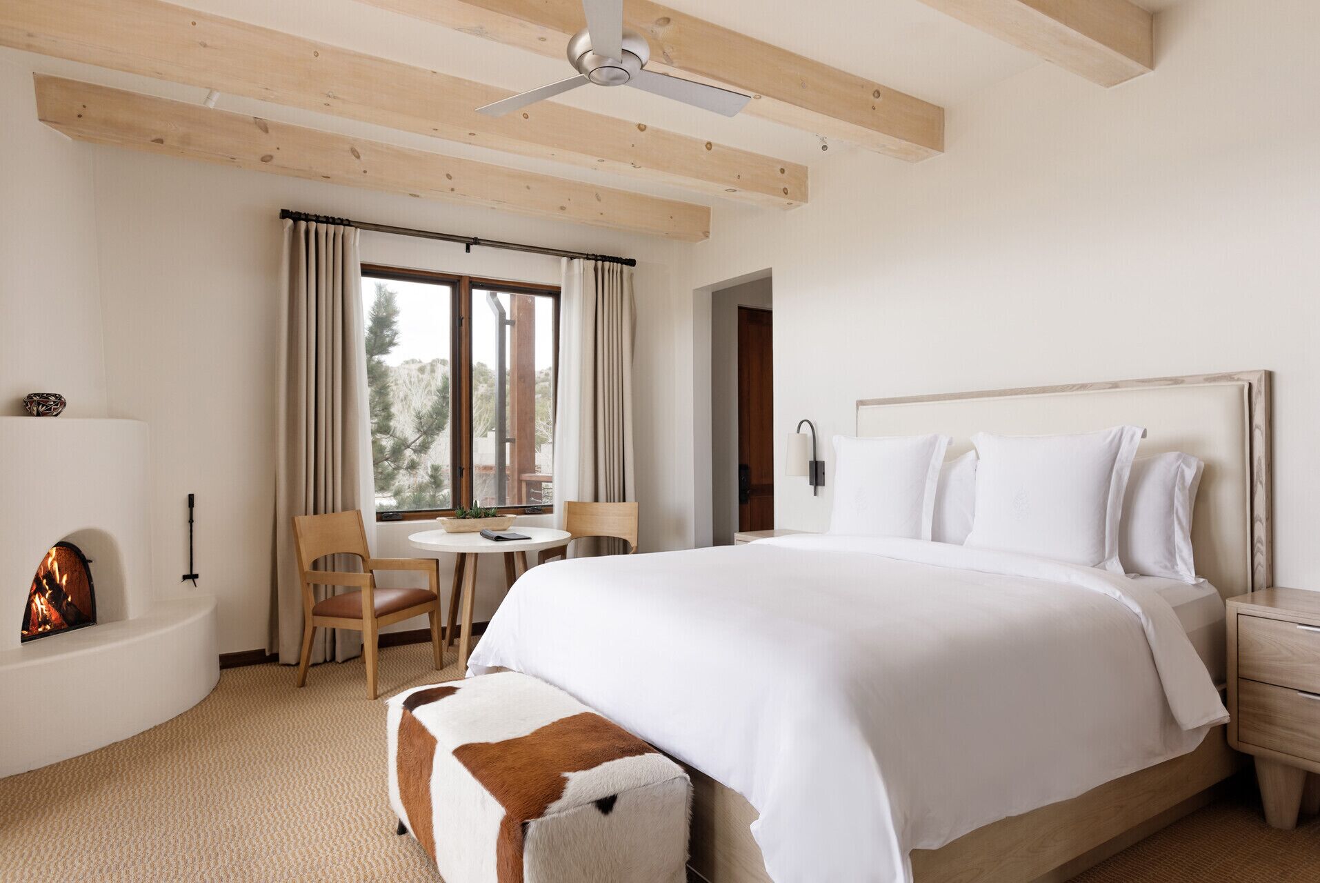 A hotel room with a large bed, a wooden ceiling, a small table with chairs, a fireplace, and a view of trees through the window.