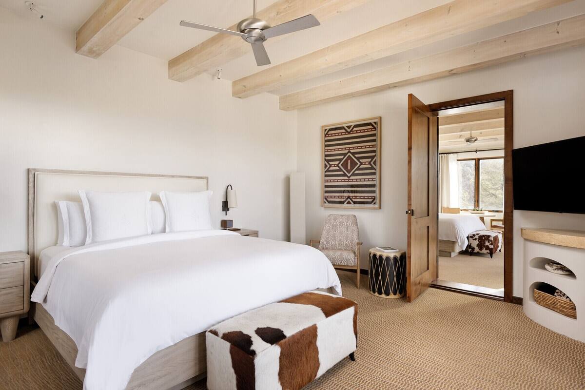 A bedroom with a large bed, a ceiling fan, a wooden ceiling, a wall-mounted TV, and a door leading to another room.