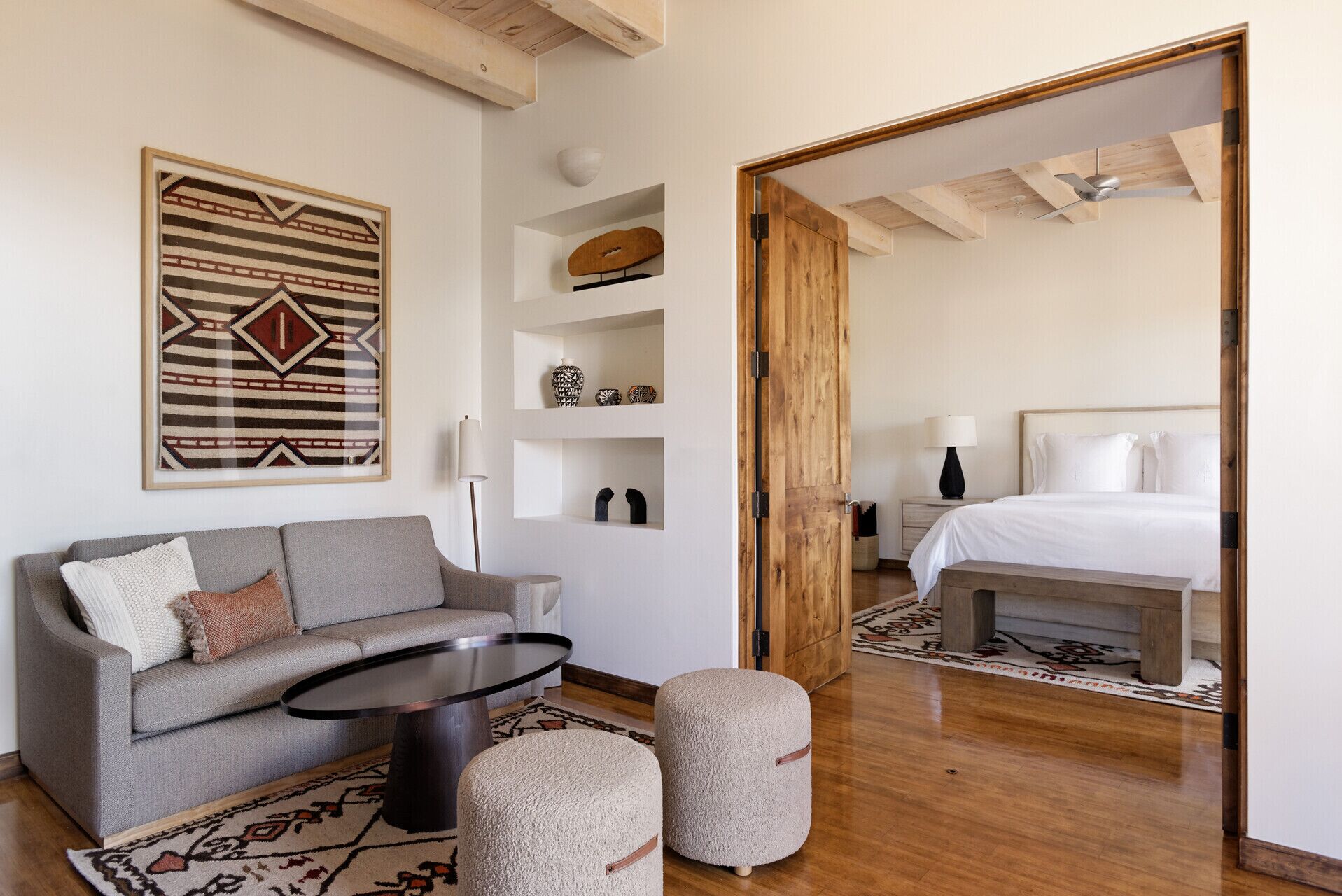 A modern room with a grey sofa, a round coffee table, and a wooden floor. There is a bed with white bedding in the adjacent room.