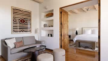 A modern room with a grey sofa, a round coffee table, and a wooden floor. There is a bed with white bedding in the adjacent room.