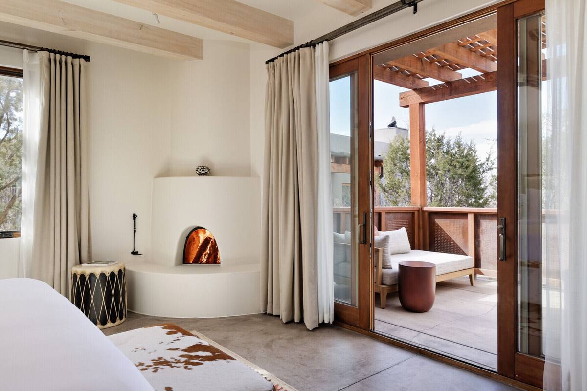 A modern bedroom with a fireplace, a large window with curtains, a wooden ceiling, and a view of the outdoors.