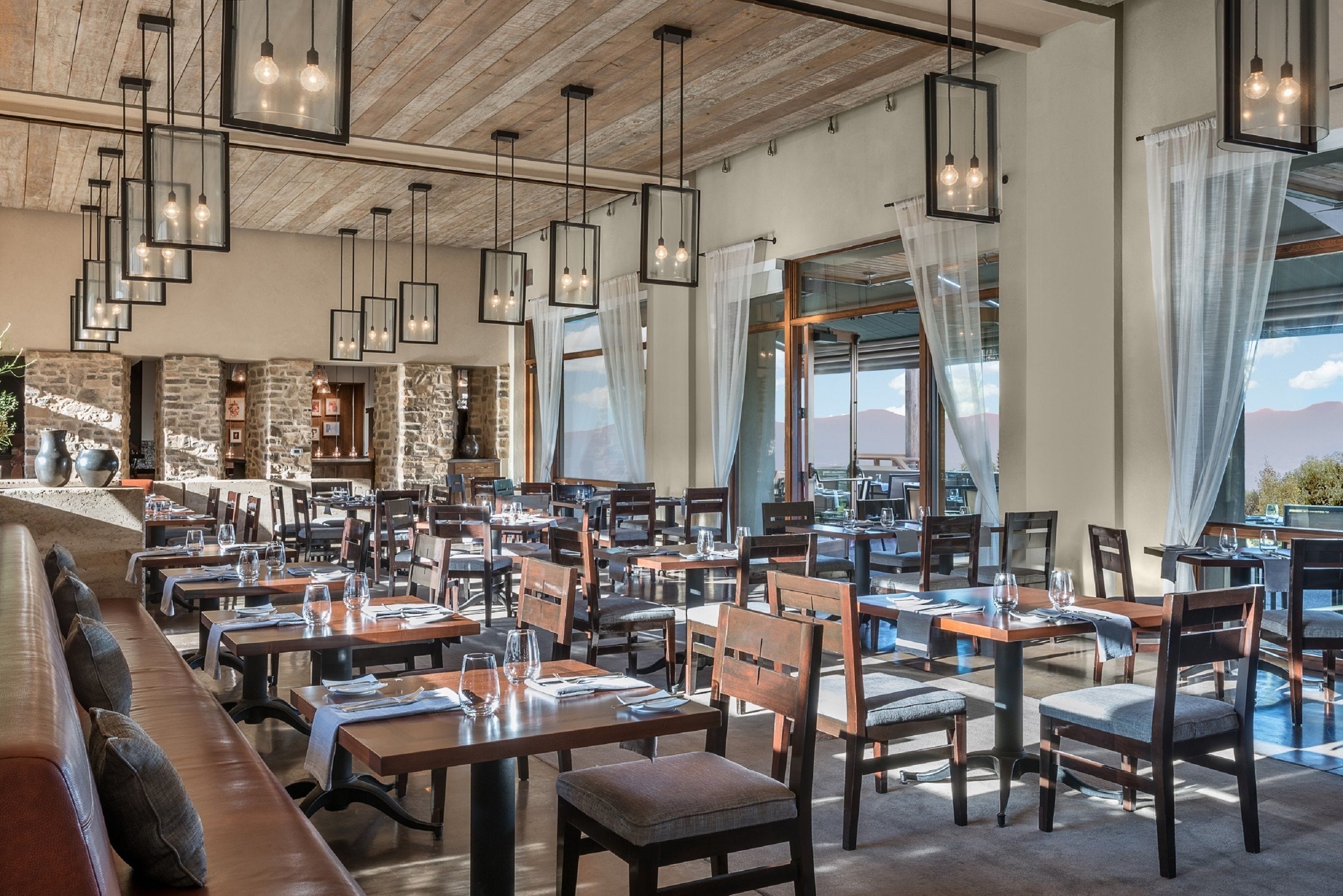 A restaurant with multiple tables and chairs, wooden beams on the ceiling, and large windows with curtains.