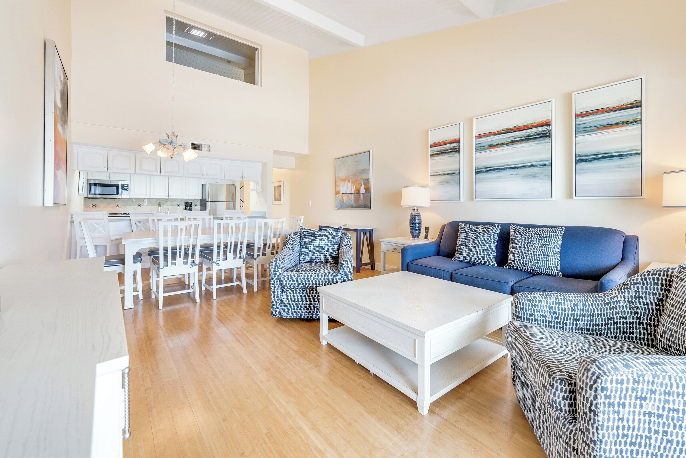 Een moderne woonkamer met een witte salontafel, een blauw gestreepte bank en bijpassende fauteuils. Op de achtergrond is een keuken met witte kasten en een eethoek met witte stoelen te zien.