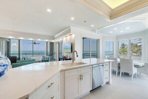A modern kitchen with a large island, a dining area with white wicker chairs, and a view of the ocean through large windows. - South Seas (Captiva)