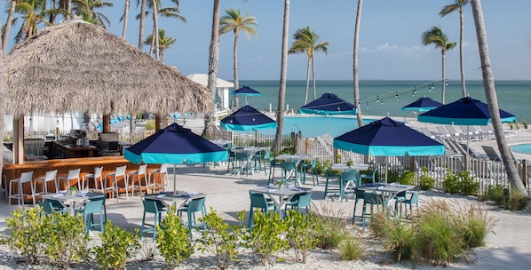A beachside restaurant with outdoor seating, a pool area, and palm trees.