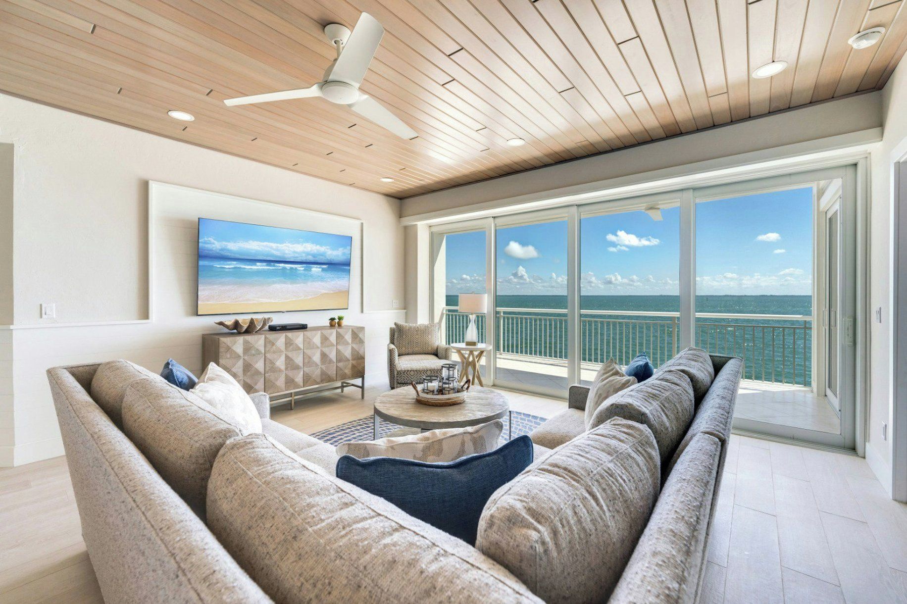 Ein geräumiges Wohnzimmer mit einem großen Flachbildfernseher, einem Deckenventilator und Meerblick durch große Fenster.