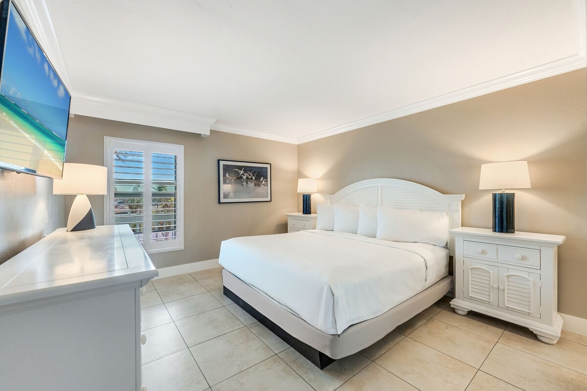 A bedroom with a bed, nightstands, a TV, and a window with blinds.