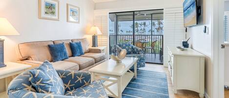 A living room with a sofa, armchair, coffee table, and a TV mounted on the wall. There is a balcony with a view of palm trees and the ocean.