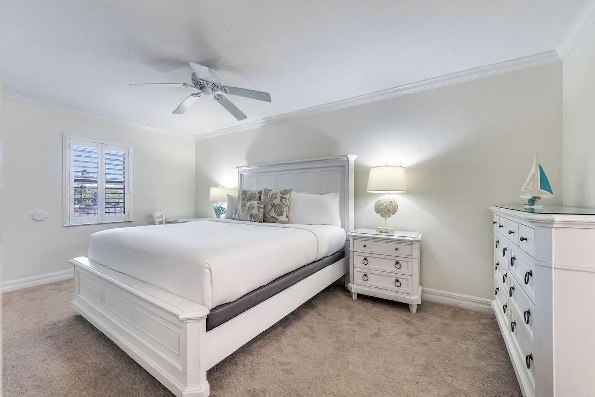 Une chambre à coucher avec un lit blanc, une table de chevet avec une lampe, une commode et un ventilateur de plafond.