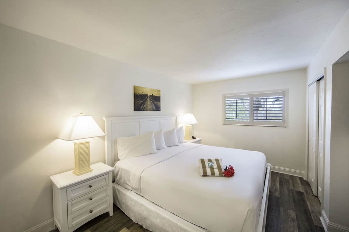 Une chambre à coucher avec un lit, deux tables de chevet avec des lampes, une fenêtre avec des stores et un tableau au mur.
