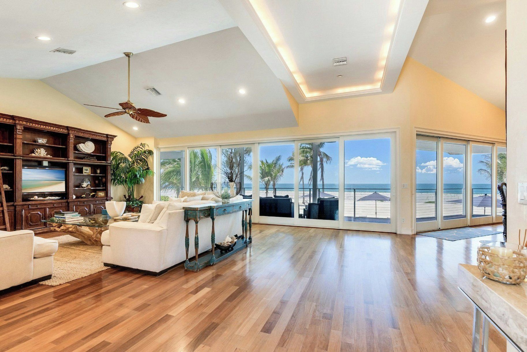 A spacious living room with a large window overlooking a beach, wooden flooring, and a ceiling fan.