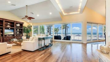A spacious living room with a large window overlooking a beach, wooden flooring, and a ceiling fan.