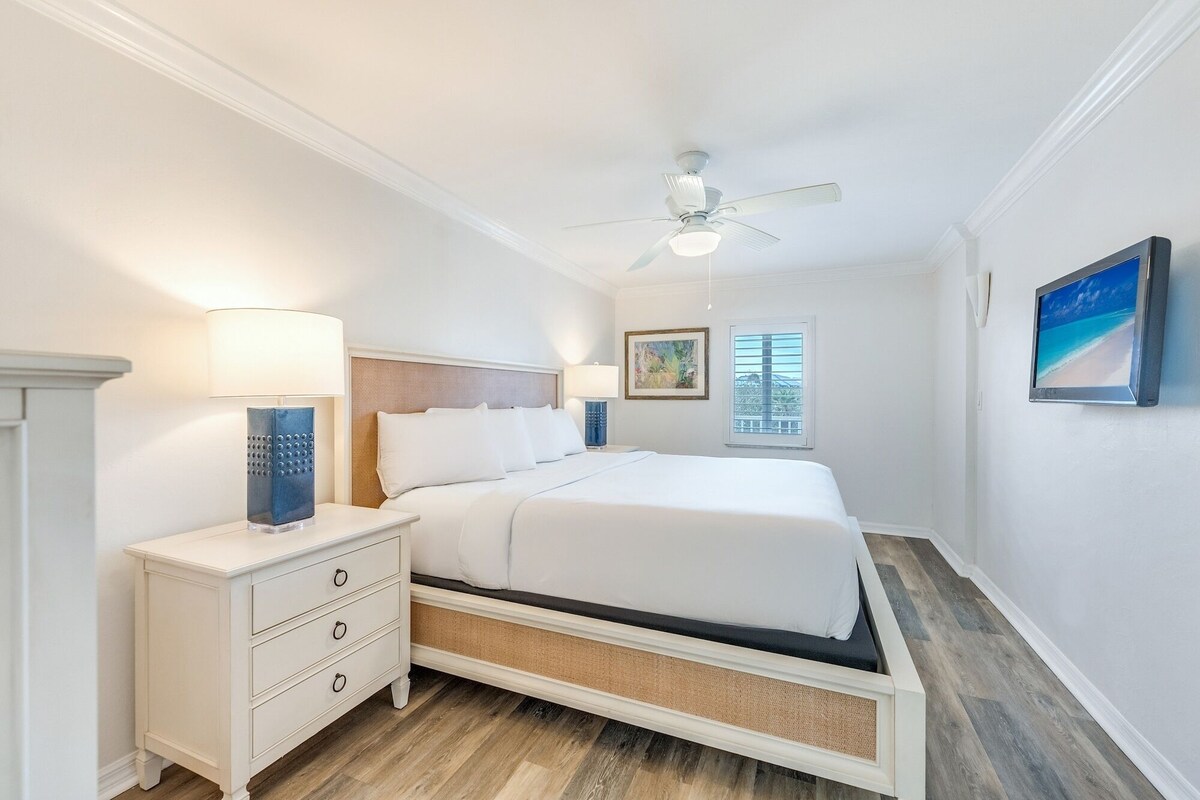 A bedroom with a bed, nightstands, a ceiling fan, a window with blinds, and a painting on the wall.