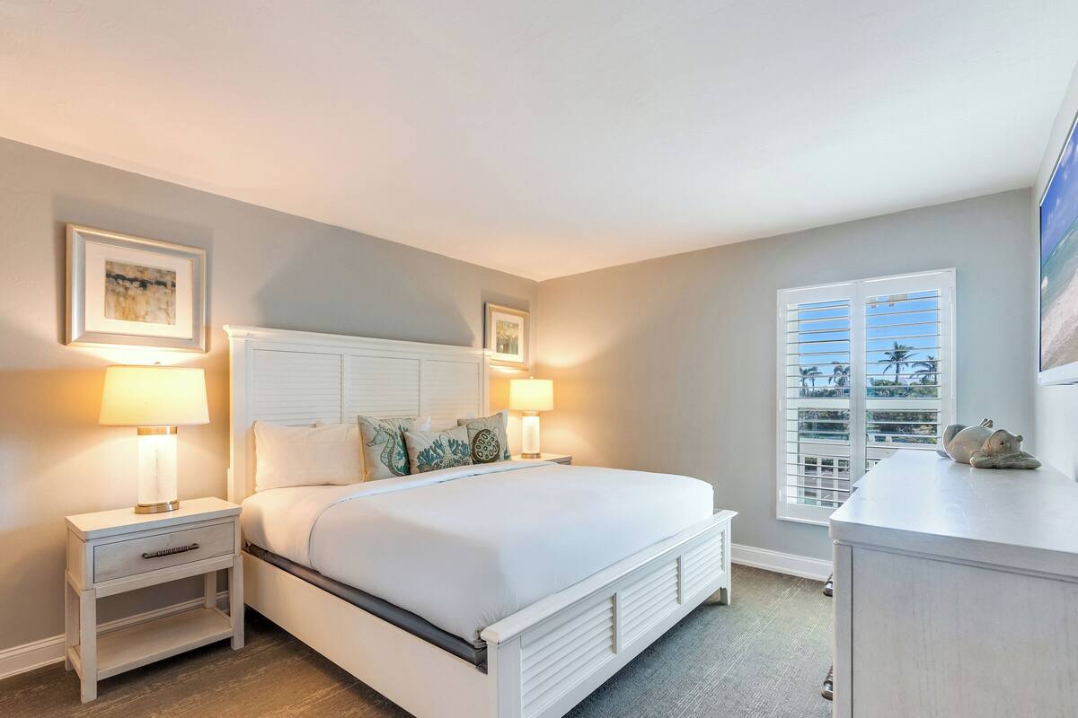 Une chambre à coucher comprenant un lit, des tables de chevet, une commode et une fenêtre avec des stores.