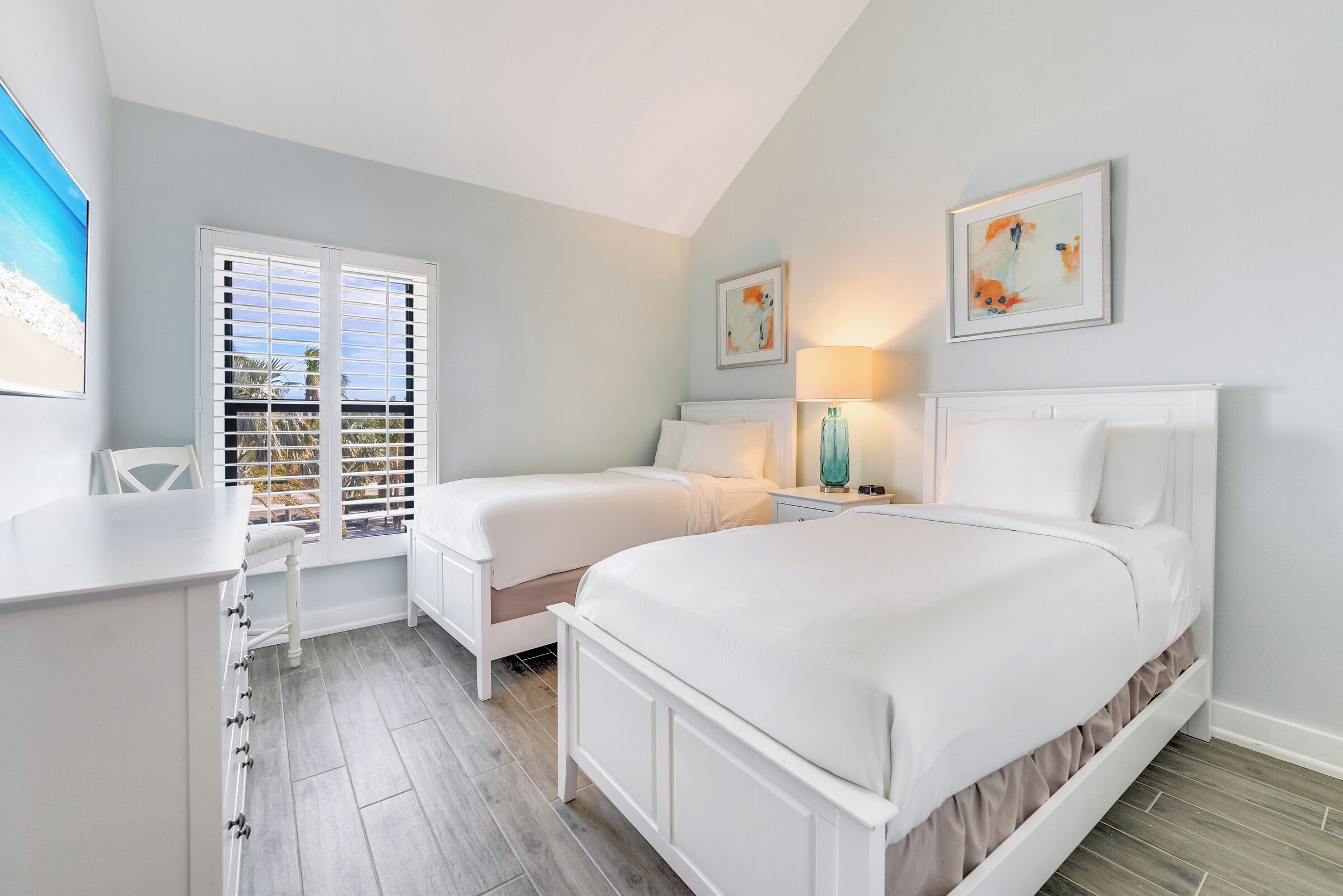 A bedroom with two beds, a desk, a chair, a window with shutters, and artwork on the walls.