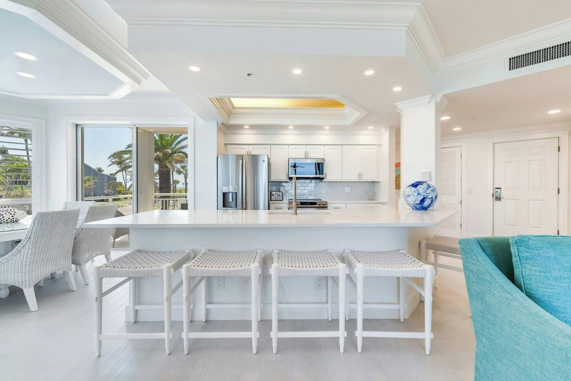 Una cocina moderna con armarios blancos, una isla central con taburetes y vistas a palmeras.