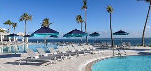 A poolside area with lounge chairs and umbrellas, palm trees, and a view of the ocean.
