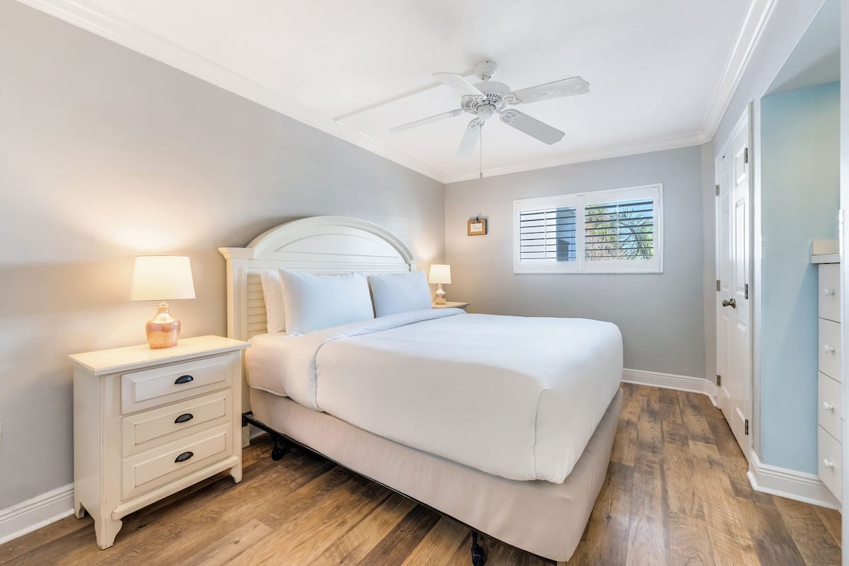 Une chambre à coucher comprenant un lit, des tables de chevet, un ventilateur de plafond, une fenêtre avec des stores et un plancher en bois.