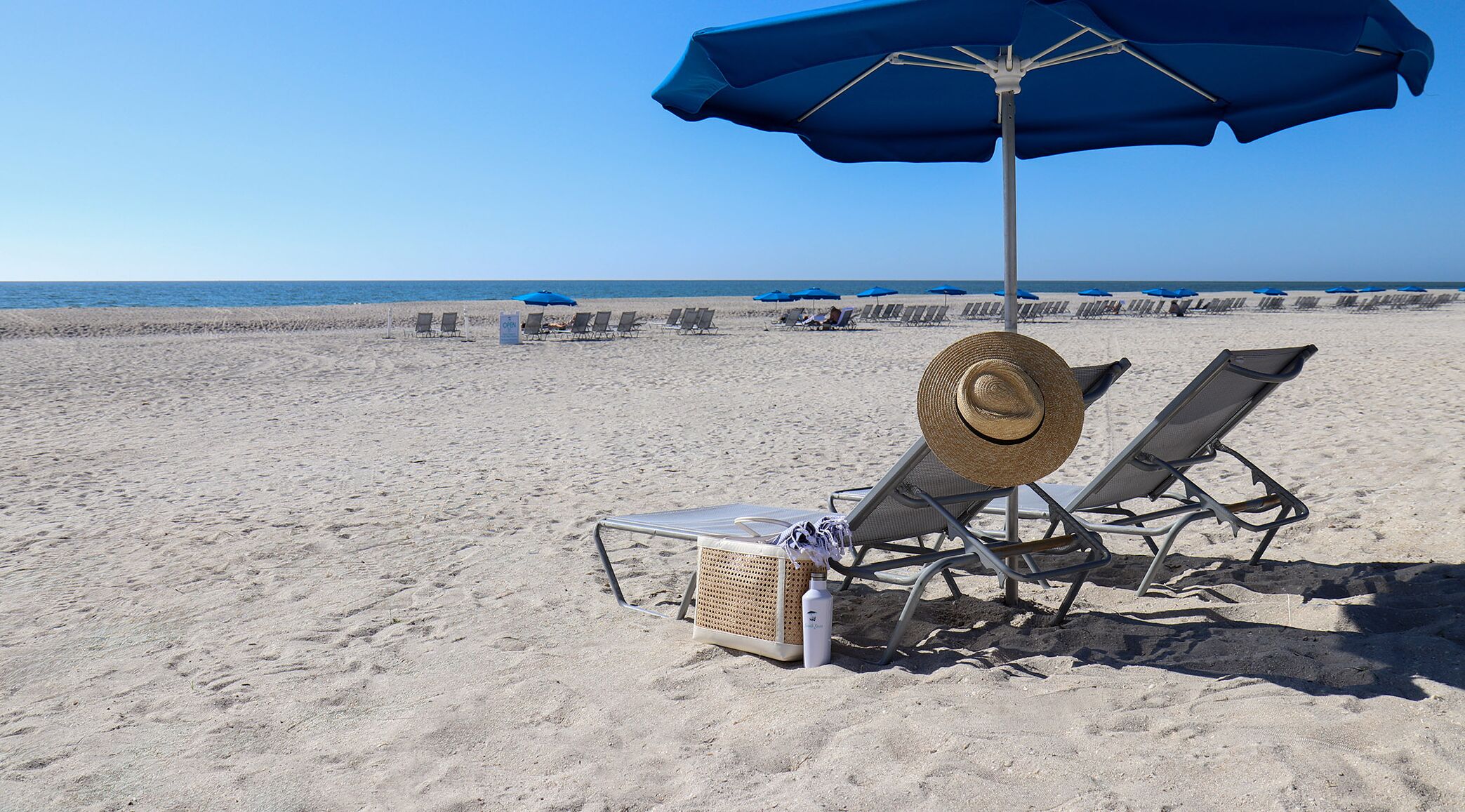 A beach with sun loungers, a straw hat, and a blue umbrella.