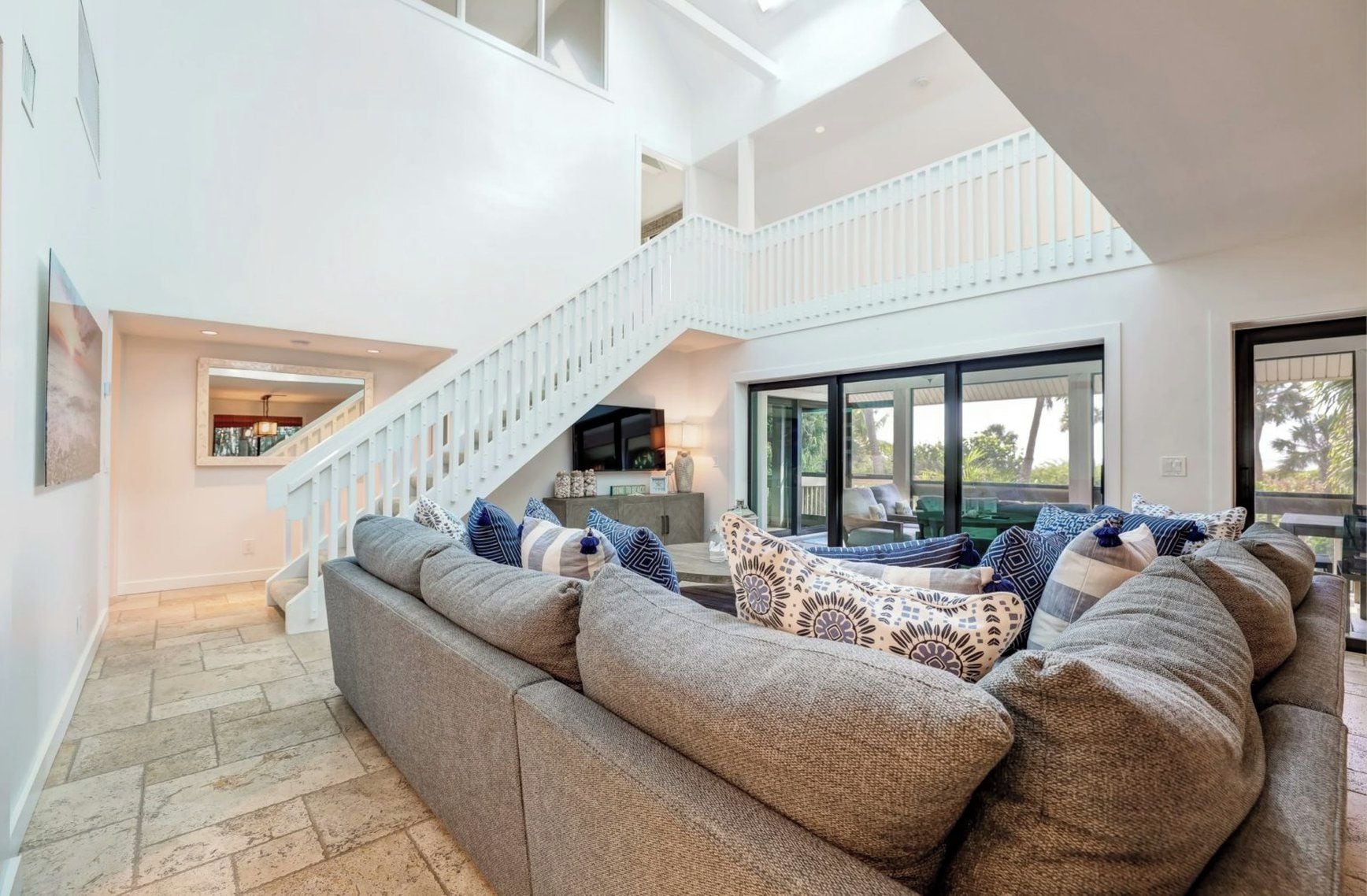 Un salón amplio con un sofá seccional gris grande, una escalera blanca y un ventanal con vistas al exterior.