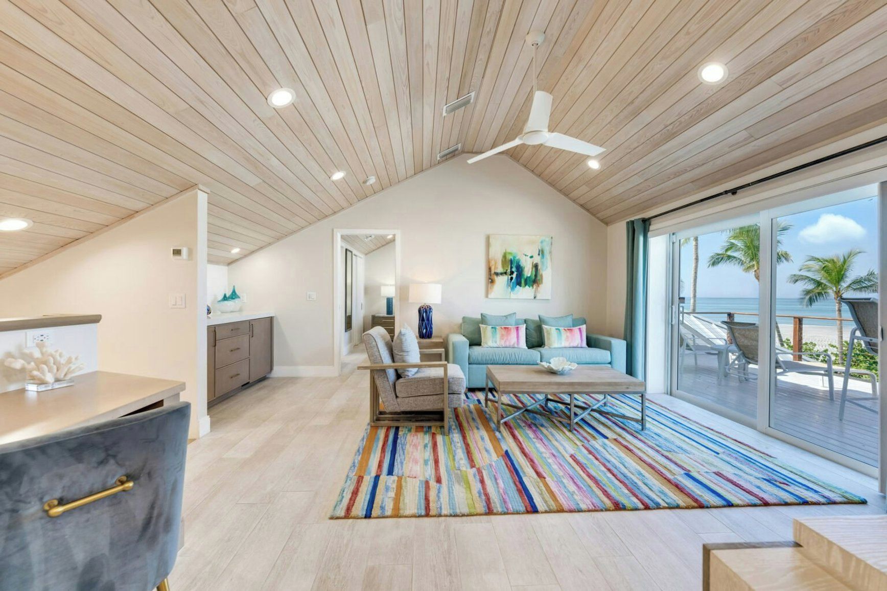 Un vaste espace de vie avec une grande fenêtre donnant sur une plage, un ventilateur de plafond et un tapis rayé aux couleurs vives.