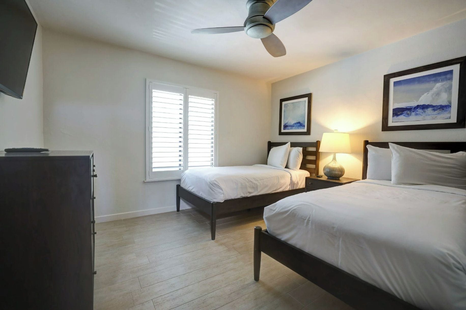 A hotel room with two beds, a ceiling fan, a desk with a lamp, and a window with blinds.