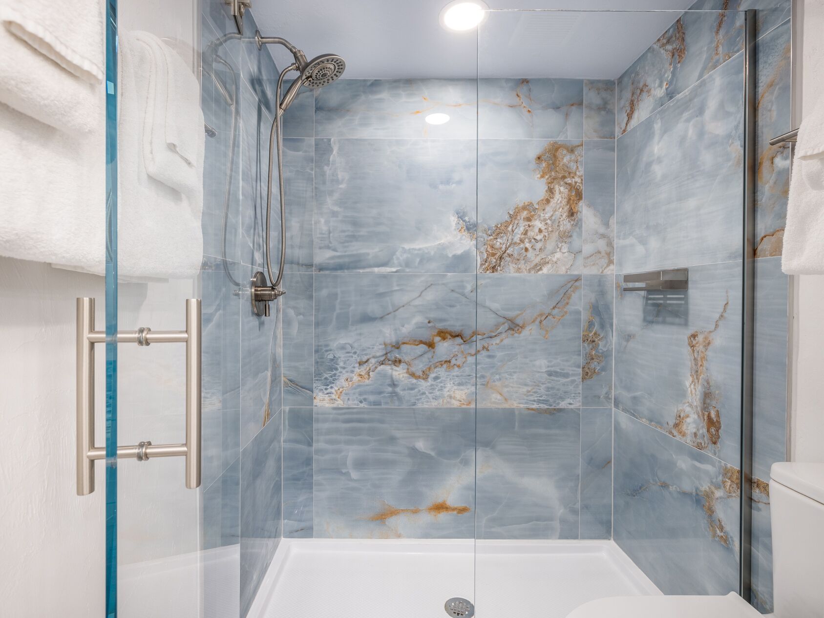 A modern shower with a glass door, marble tiles, and a handheld showerhead.