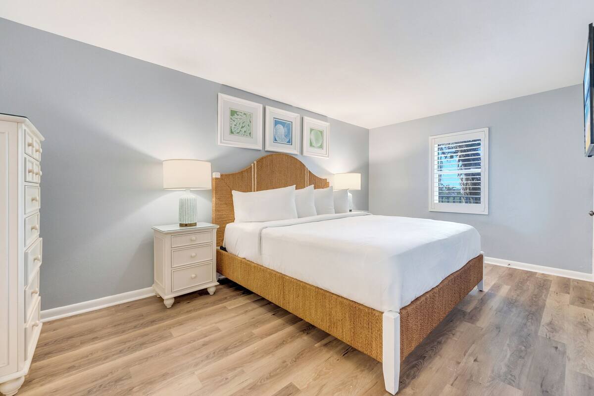 Une chambre à coucher avec un lit doté d’une tête de lit en bois, une table de chevet avec une lampe, une commode et une fenêtre avec des stores.