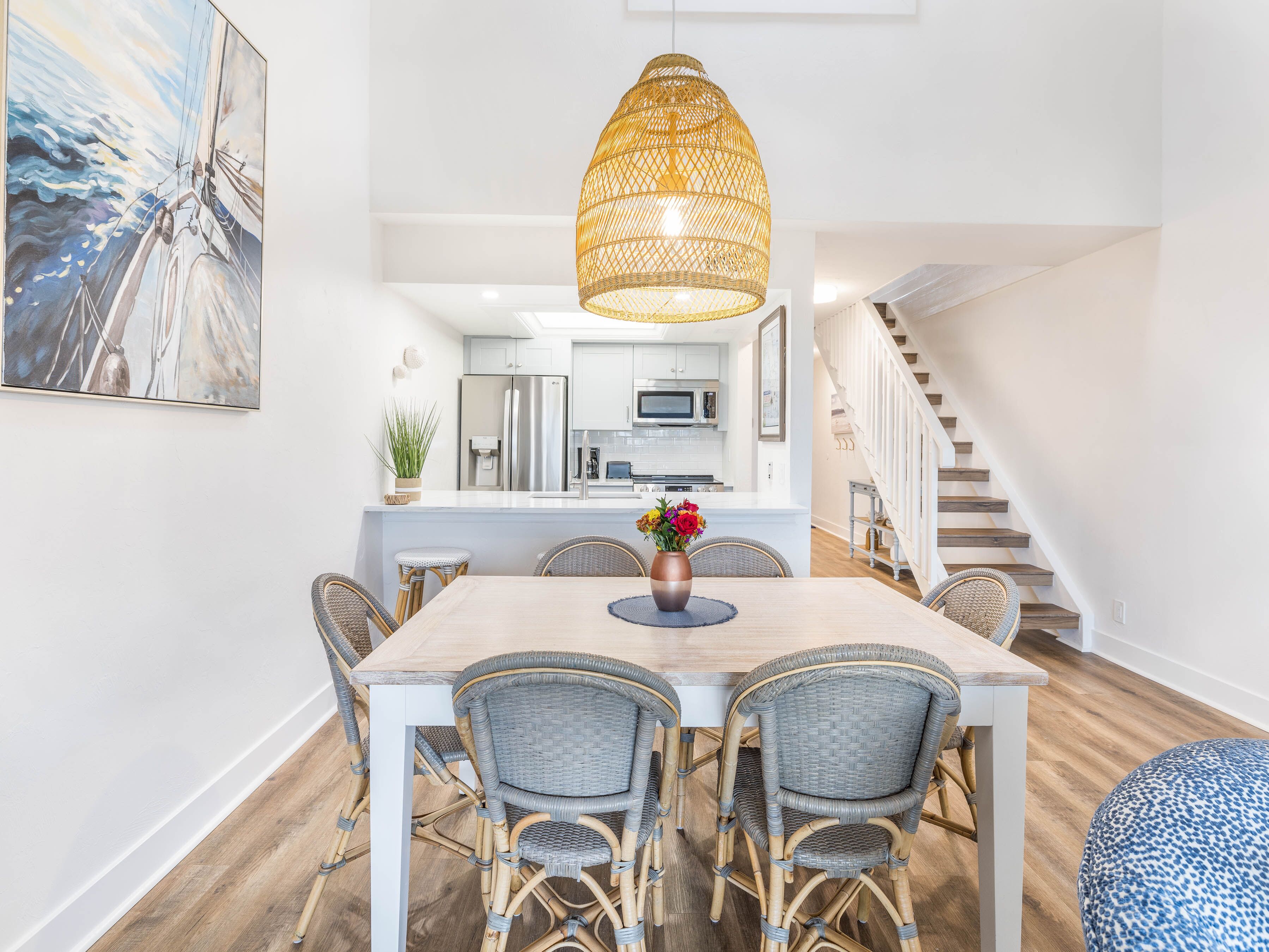 A dining area with a wooden table, wicker chairs, a hanging light fixture, and a kitchen in the background.