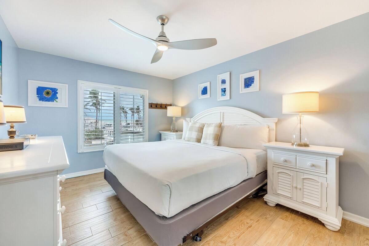 Une chambre à coucher comprenant un lit, une table de chevet, une commode, un ventilateur de plafond et une fenêtre avec des stores.