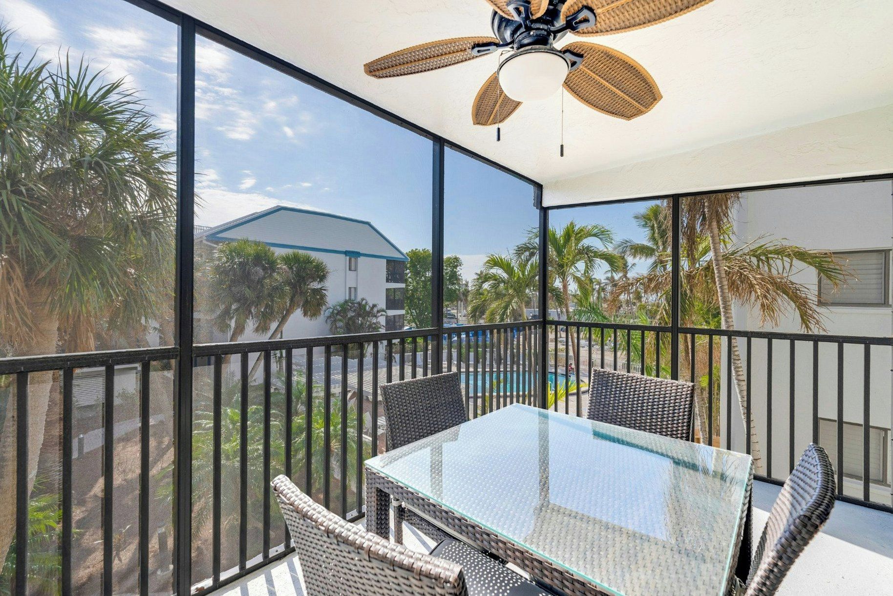 A balcony with a glass table and wicker chairs, overlooking a pool and palm trees.