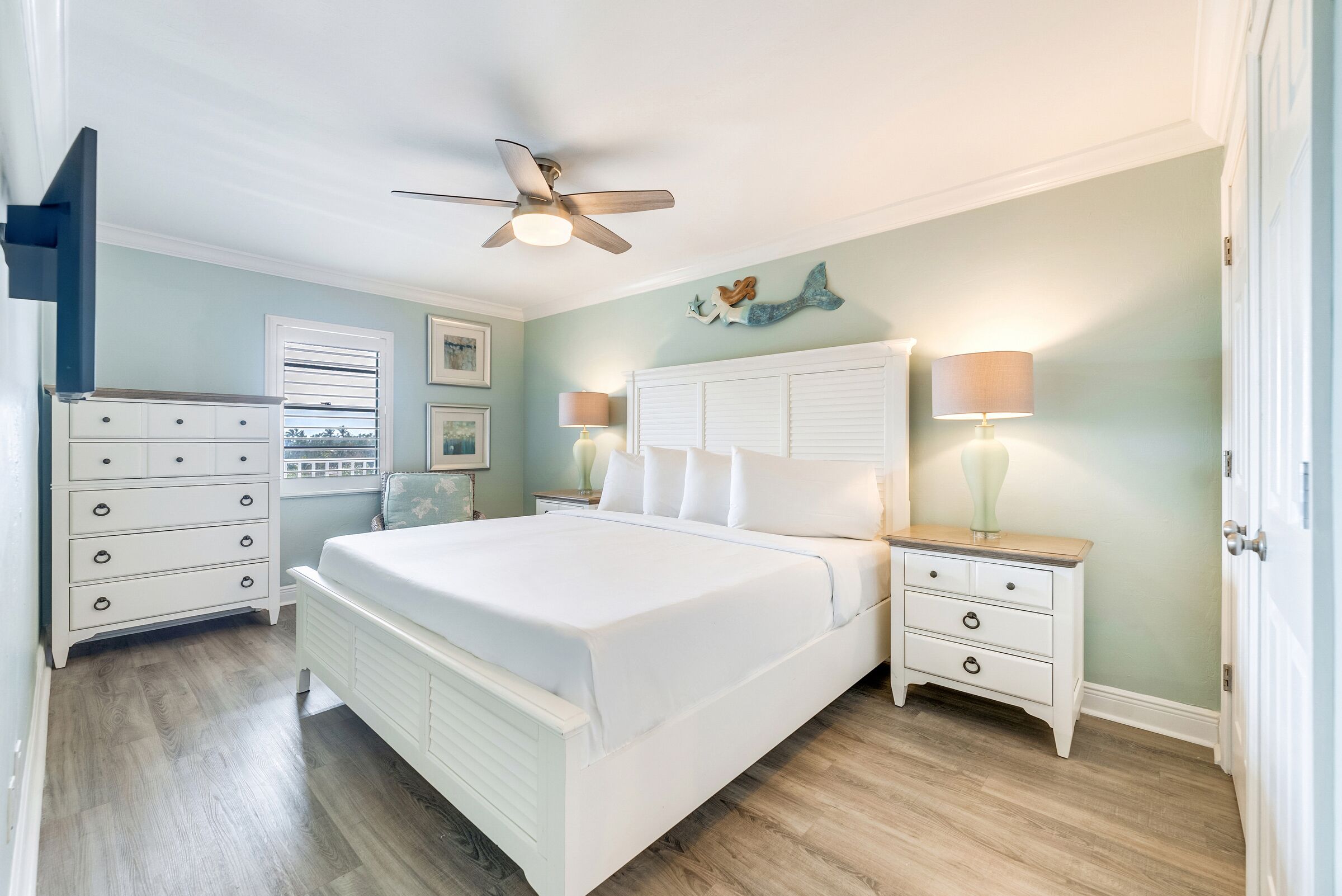 A bedroom with a large bed, white nightstands, a ceiling fan, and a window with blinds.