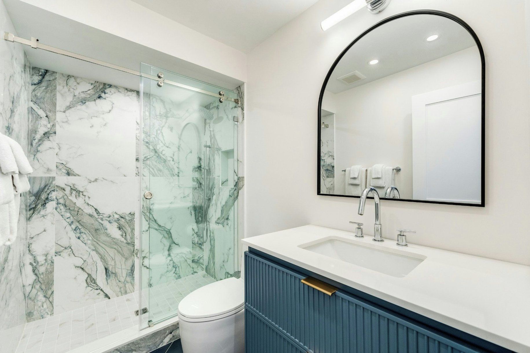 A bathroom with a glass shower enclosure, a white sink with a blue cabinet, and a large mirror.