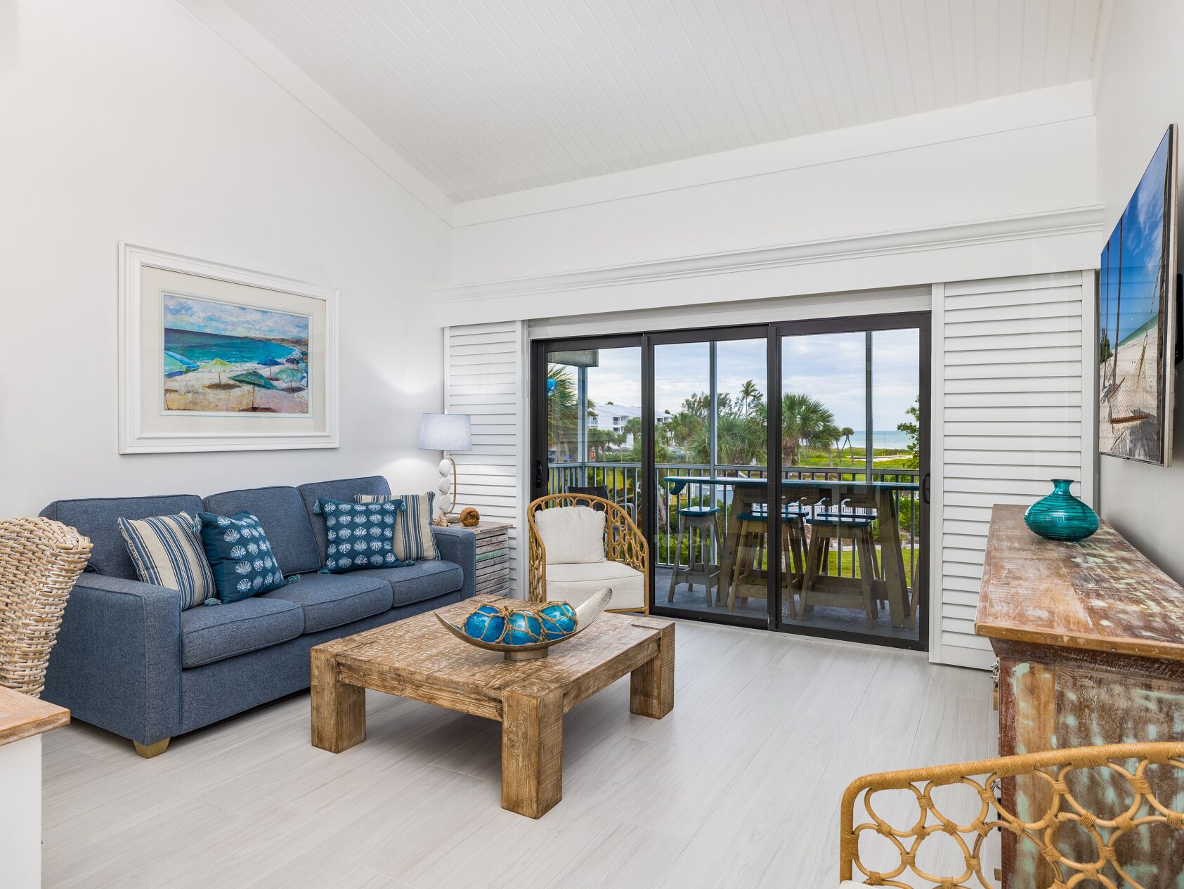 A living room with a sofa, coffee table, and wicker chairs. There is a large window with a view of the ocean and palm trees.