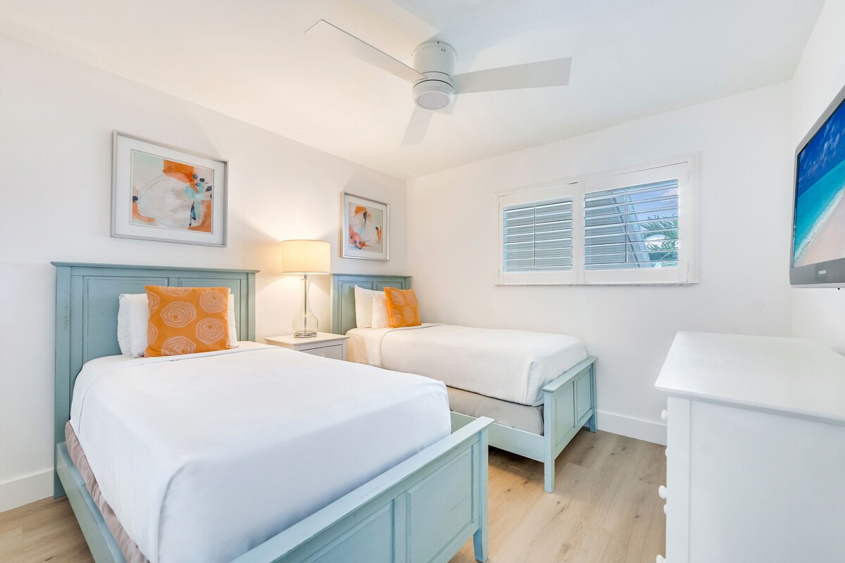 A bedroom with two beds, a ceiling fan, artwork on the walls, a window with blinds, and a TV on the right.
