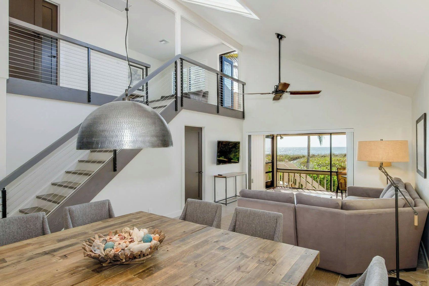 A spacious living area with a high ceiling, a staircase, a dining table with a decorative centerpiece, a sofa set, and a view of the outdoors.