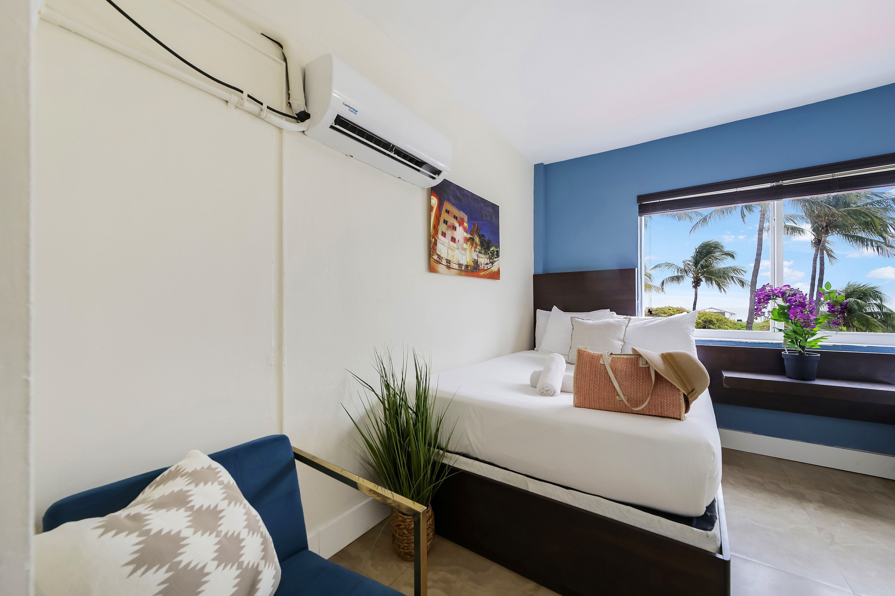Habitación de hotel con cama, sofá azul, planta en maceta, ventana con vista a palmeras y un equipo de aire acondicionado.