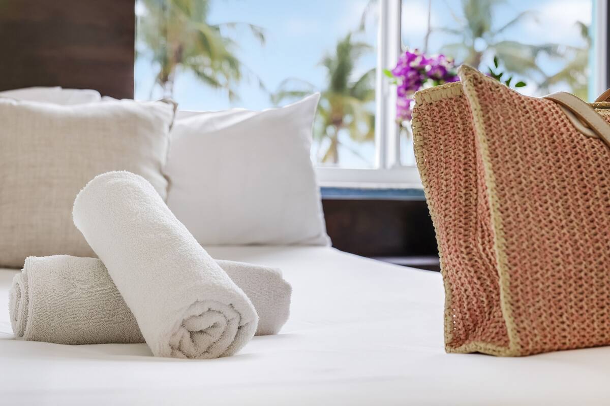 Un letto rifatto con asciugamani bianchi arrotolati e una borsa rosa con texture, probabilmente per gli ospiti, con vista su palme e una finestra.