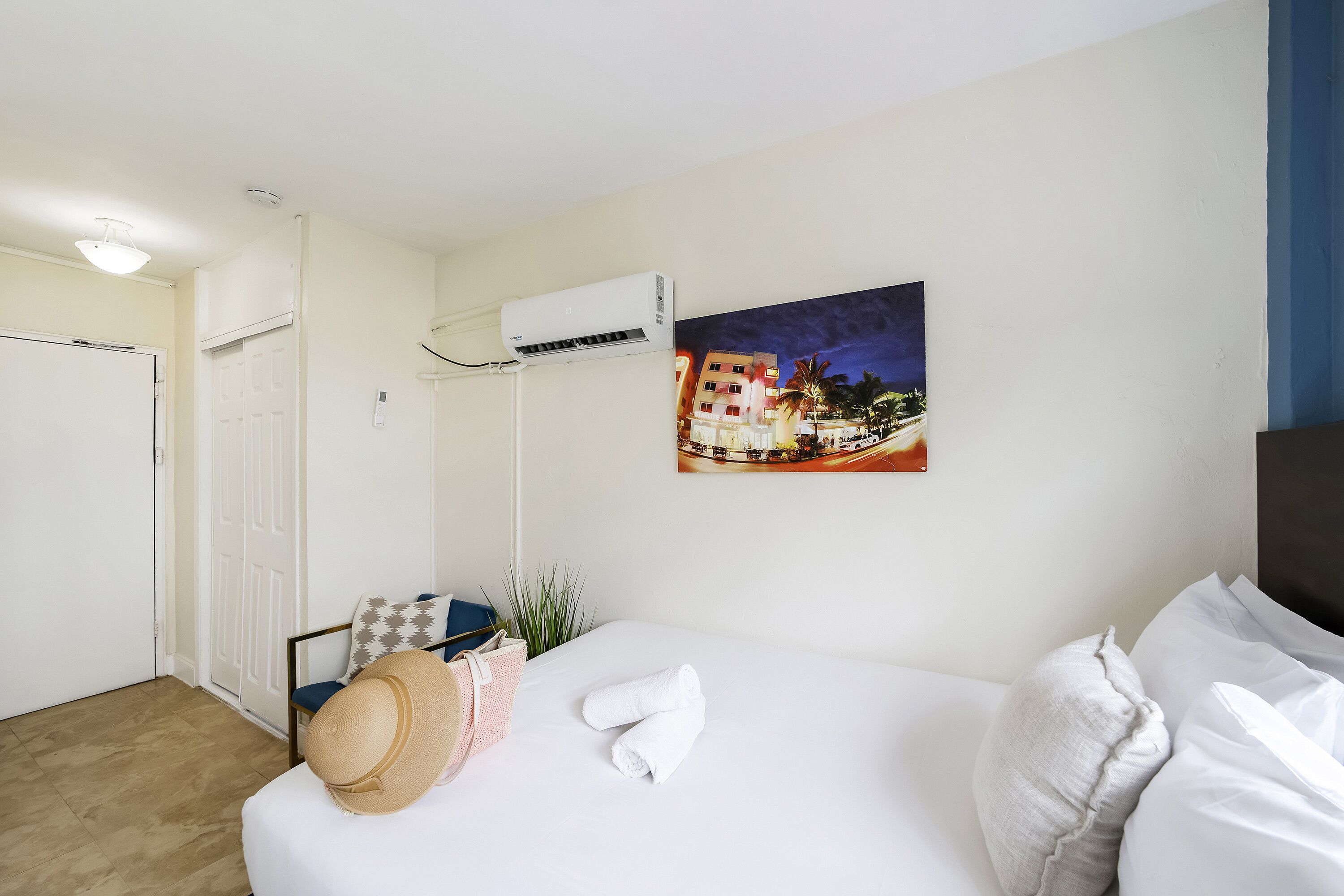 A hotel room with a bed, a painting of a cityscape, an air conditioning unit, and a door.