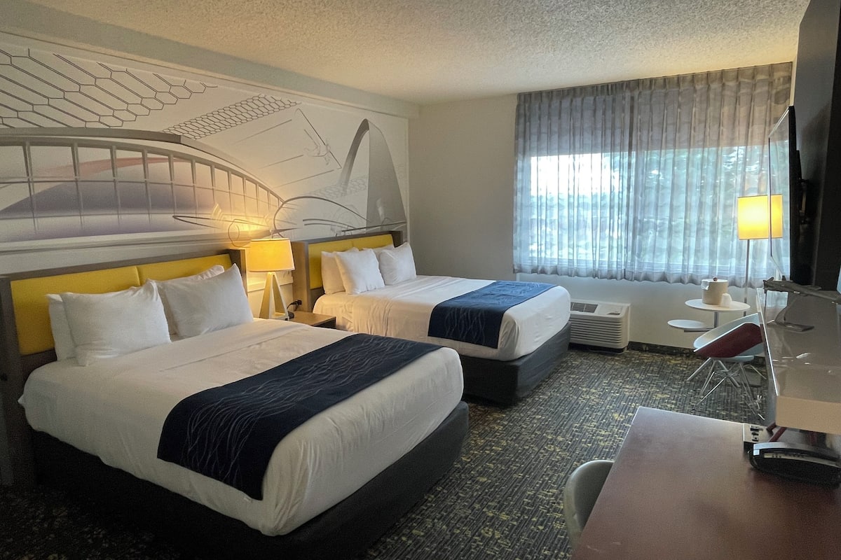 A hotel room with two beds, a desk, a chair, a lamp, and a large window with curtains.