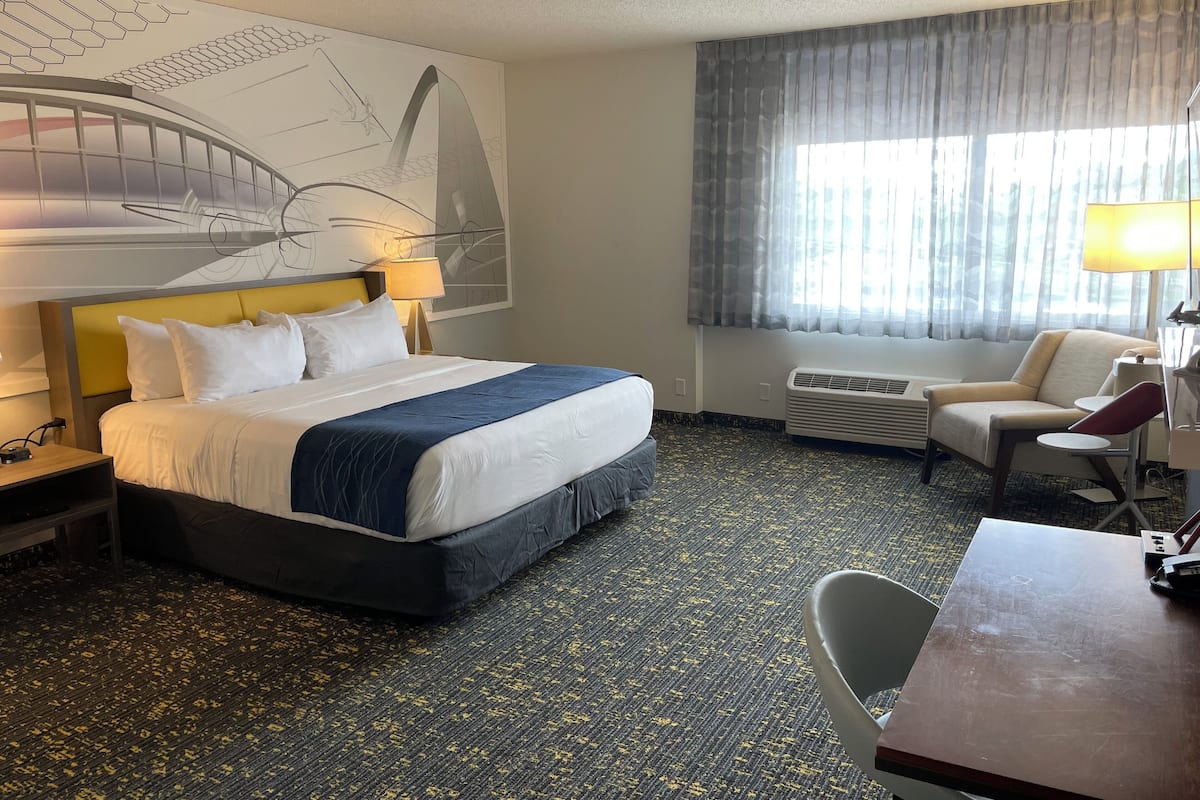 A hotel room with a large bed, a desk, a chair, a large window with curtains, a wall art piece, and an air conditioning unit.