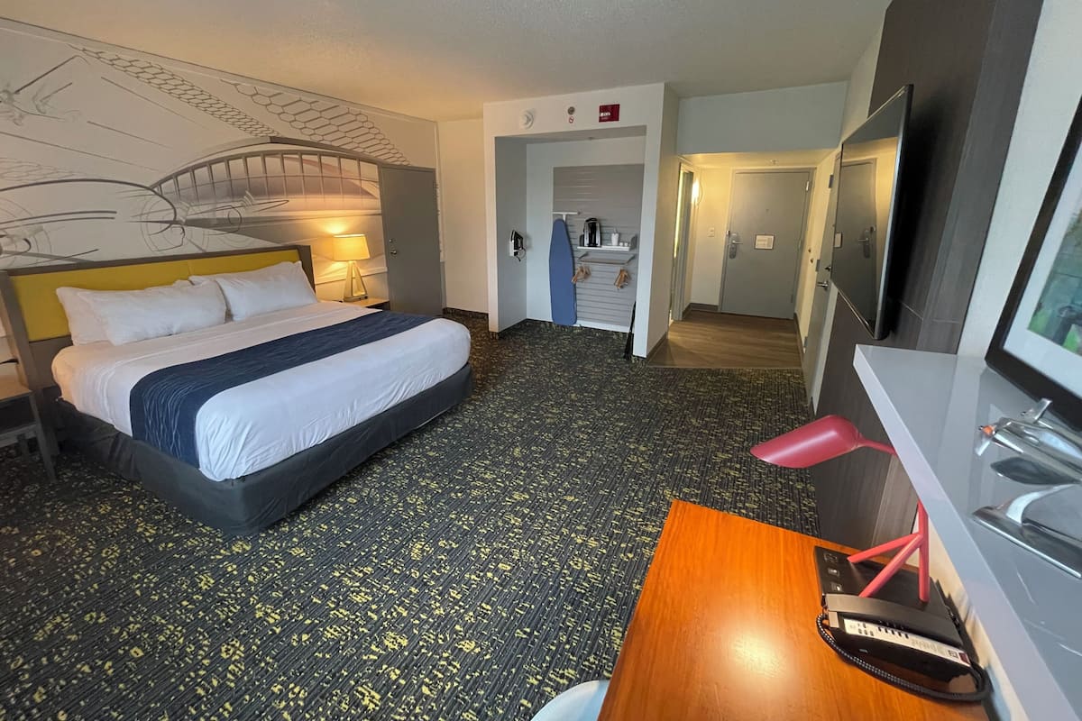 A hotel room with a bed, a desk with a computer, a TV, and a bathroom visible through an open door.