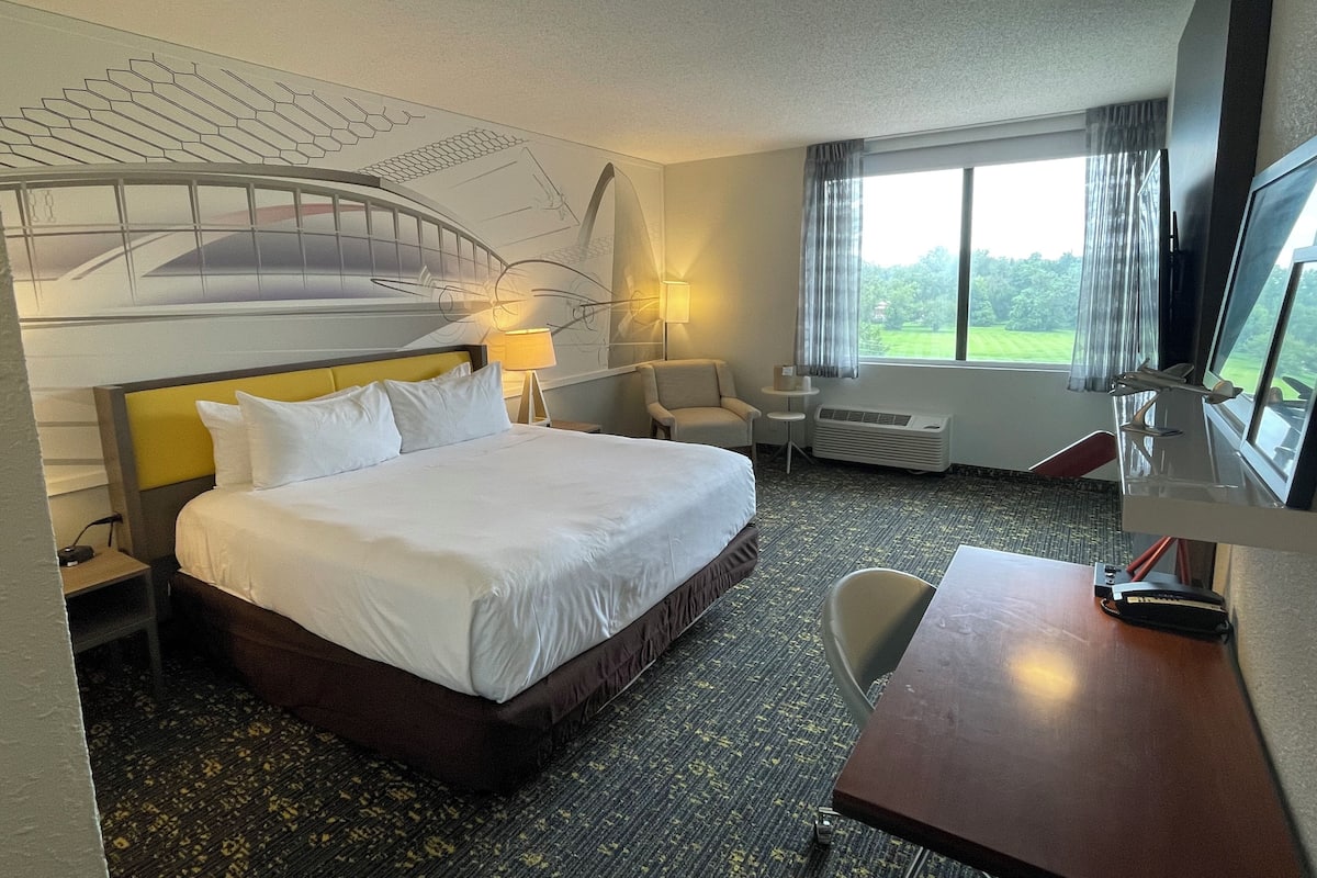 A hotel room with a large bed, a desk with a computer, a chair, a TV, and a window with a view of greenery.