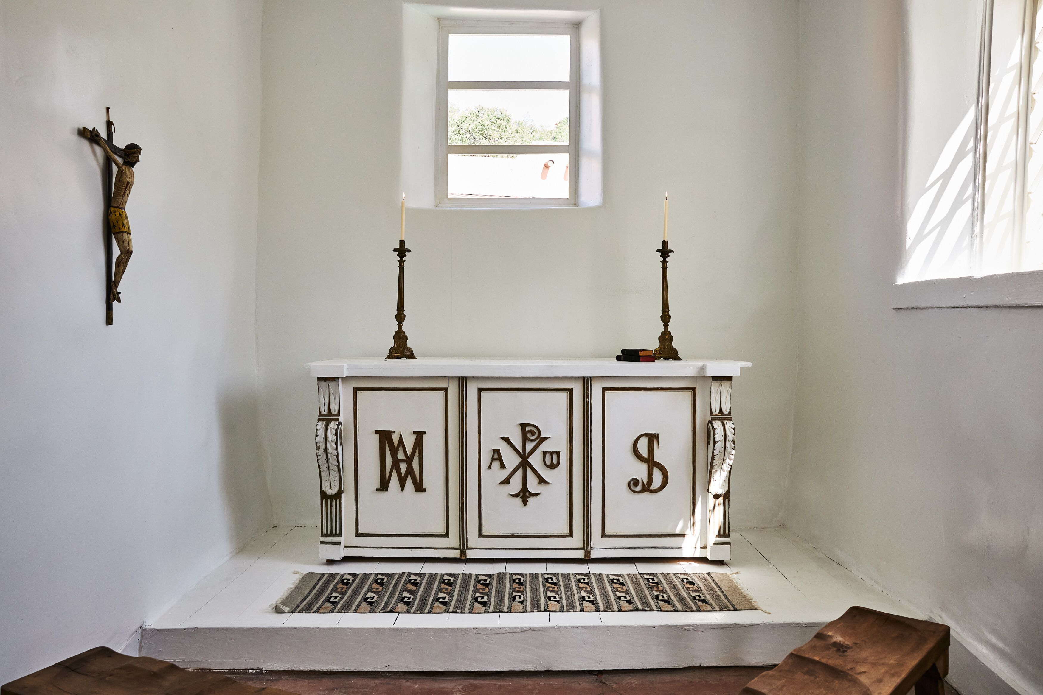 A white altar with three cabinets, each with a letter. There is a crucifix on the wall and a window above the altar.