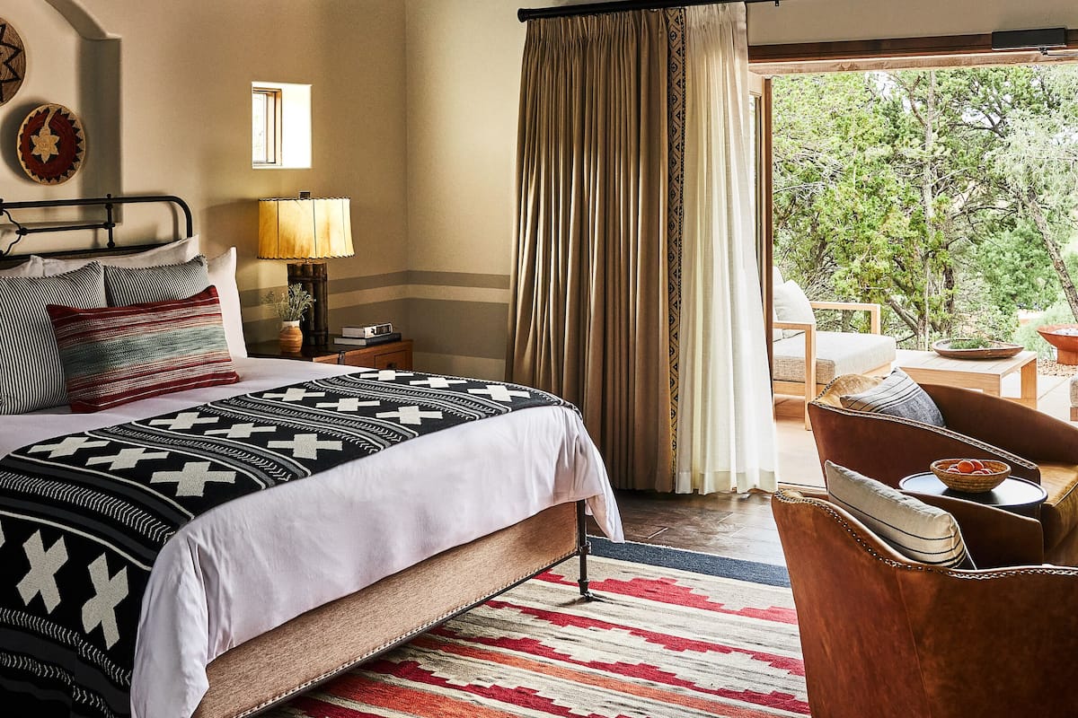A bedroom with a bed, a nightstand, a lamp, a window with curtains, and a view of the outdoors.