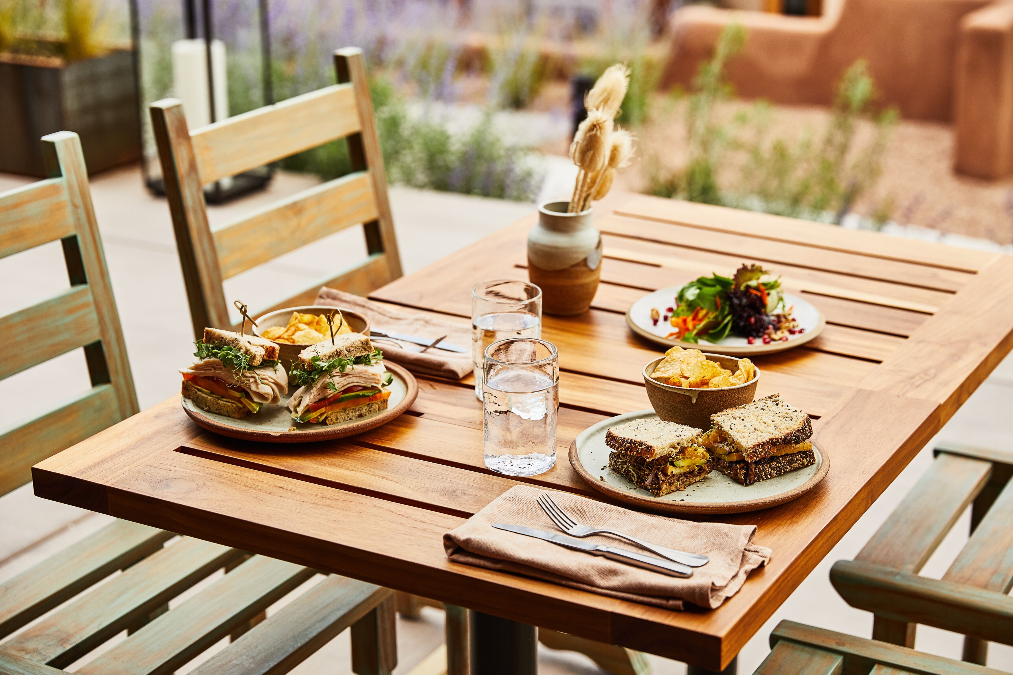 A wooden table with plates of sandwiches, a bowl of chips, and a glass of water.