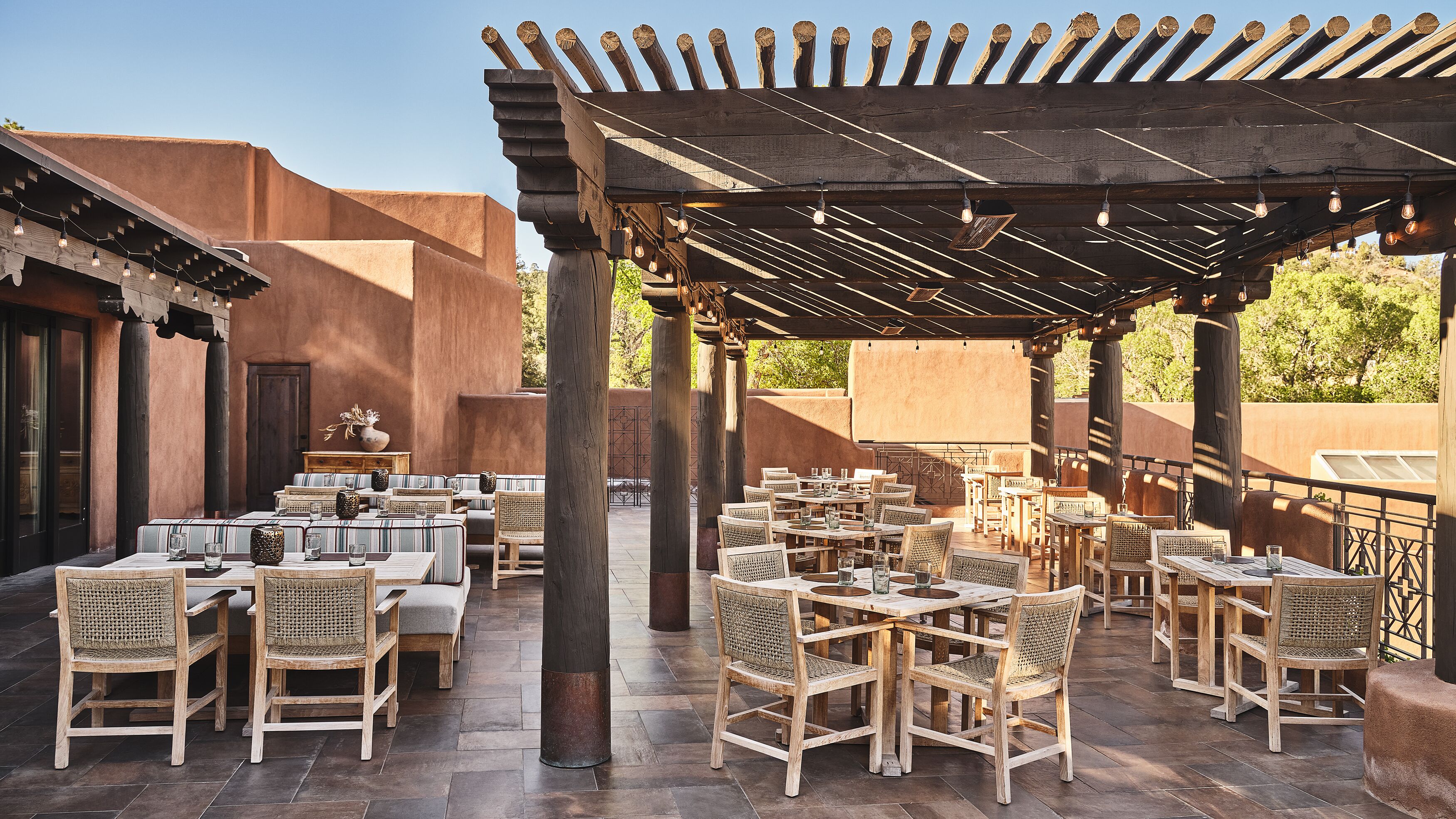 An outdoor patio with wooden tables and wicker chairs, a pergola overhead, and a rustic building in the background.